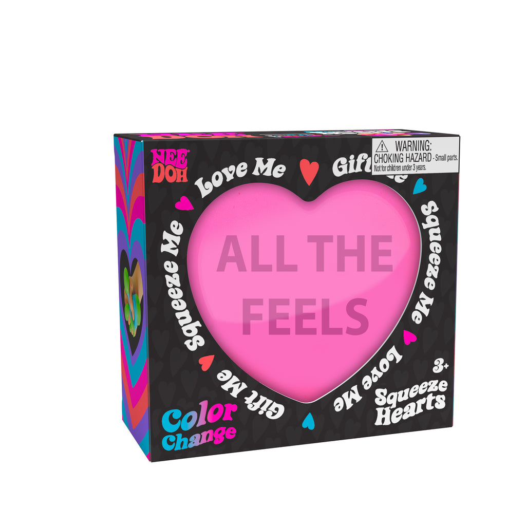 A boxed Nee Doh Color Change Squeeze Heart toy. The packaging is predominantly black with colorful accents, featuring phrases like "ALL THE FEELS," "Squeeze Me," "Love Me," and "Gift Me." The box also includes a choking hazard warning for small parts.