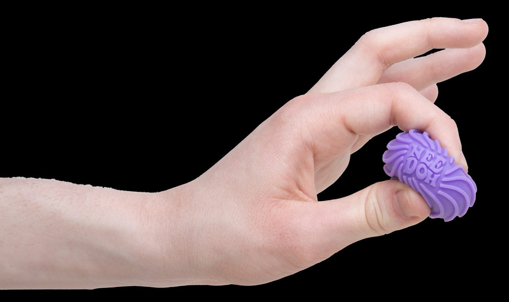 A hand is squeezing a small, purple, textured ball between two fingers. The ball's surface features wavy ridges and is called a Teenie NeeDoh Ripples. The background is plain white.